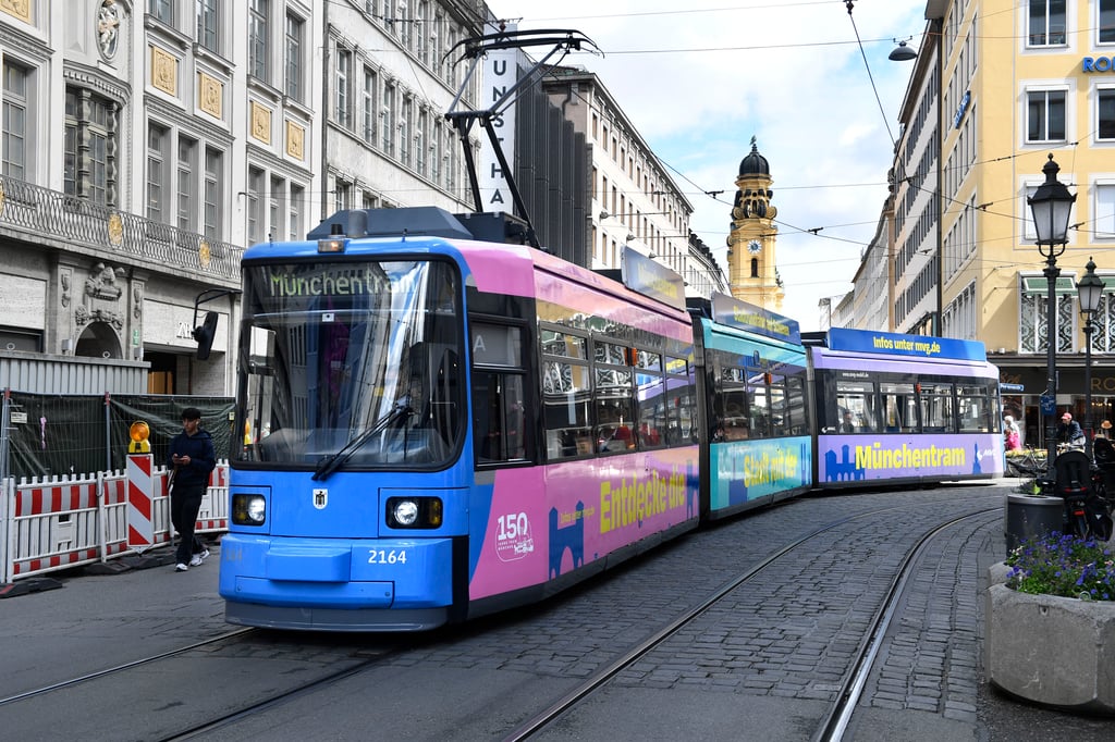 Blue and pink tram on a cobblestone street, with historic buildings and a church clock tower in the background.