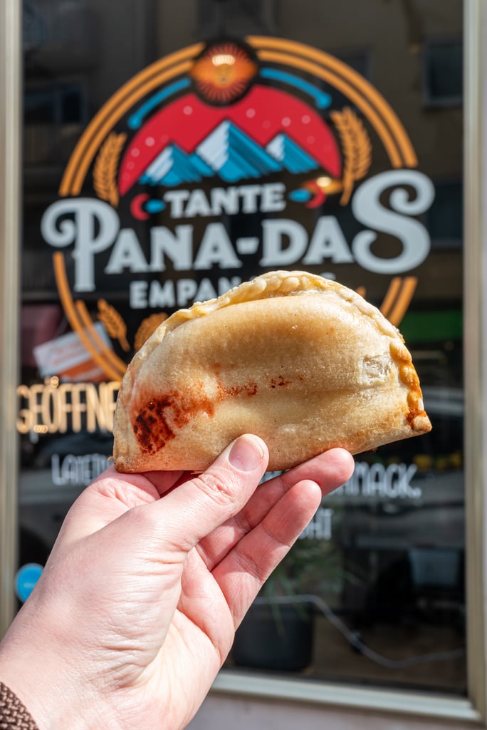 Hand holding a baked empanada in front of a storefront window with a colorful sign in the background (TANTE PANADAS).