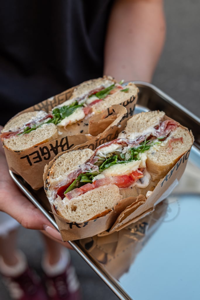 Two halves of a sandwich filled with greens, tomato, and cheese on rustic bread, held over a tray in paper wrap.