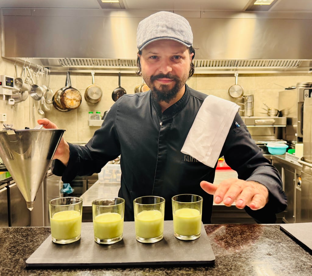 Chef in a uniform and gray cap presents five green smoothies lined up on a counter in a professional kitchen.