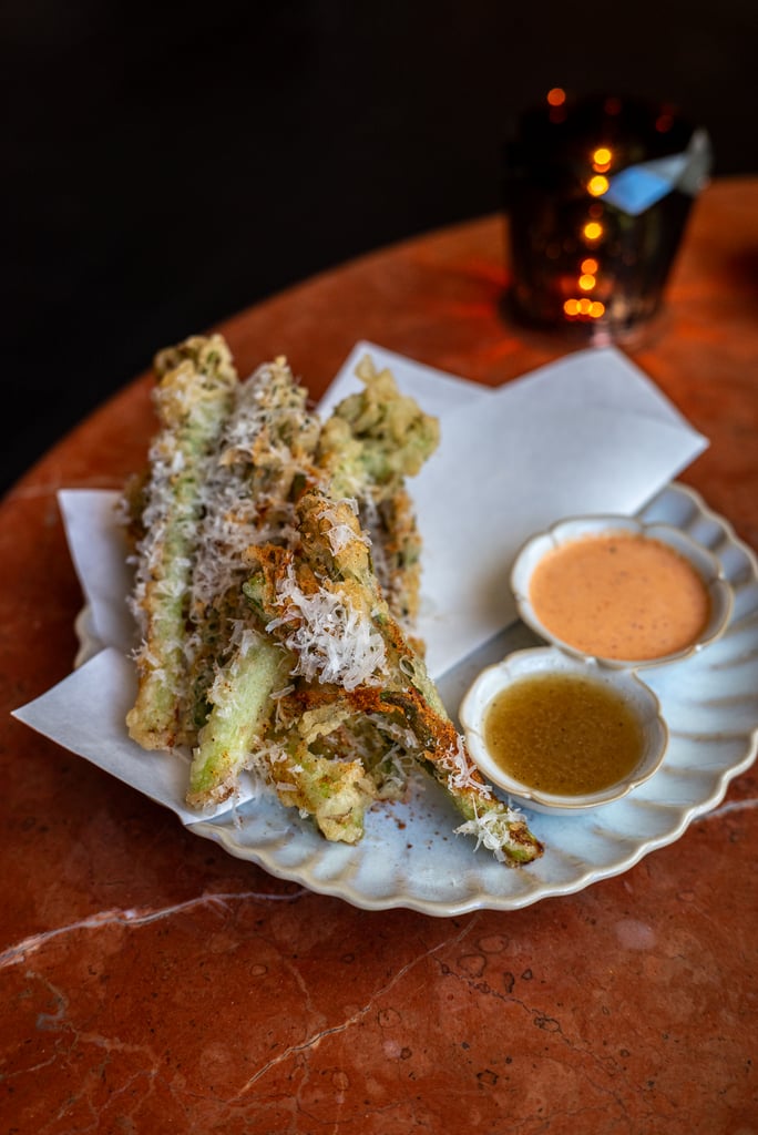 Stack of crispy tempura green beans spears with cheese flakes, served on a napkin with three dipping sauces on a scalloped plate.