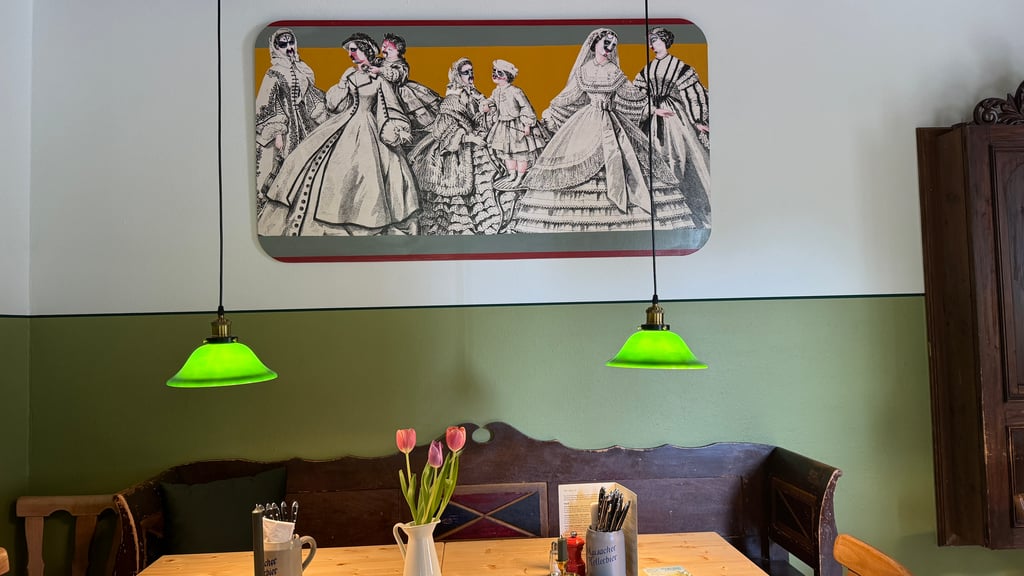 Cozy dining area with a vintage black-and-white mural of people in 19th-century dress on an olive-green wall, and two green pendant lights over a wooden table with a tulip centerpiece.