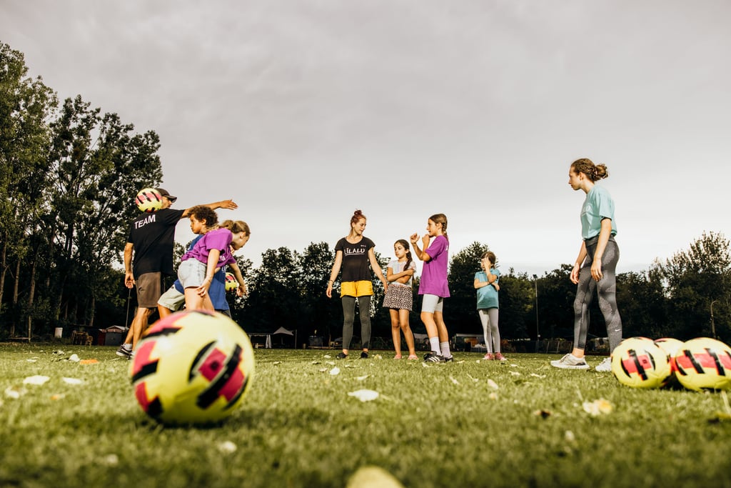Group of children standing on a grassy field during soccer practice, with colorful balls scattered nearby and a coach guiding them.