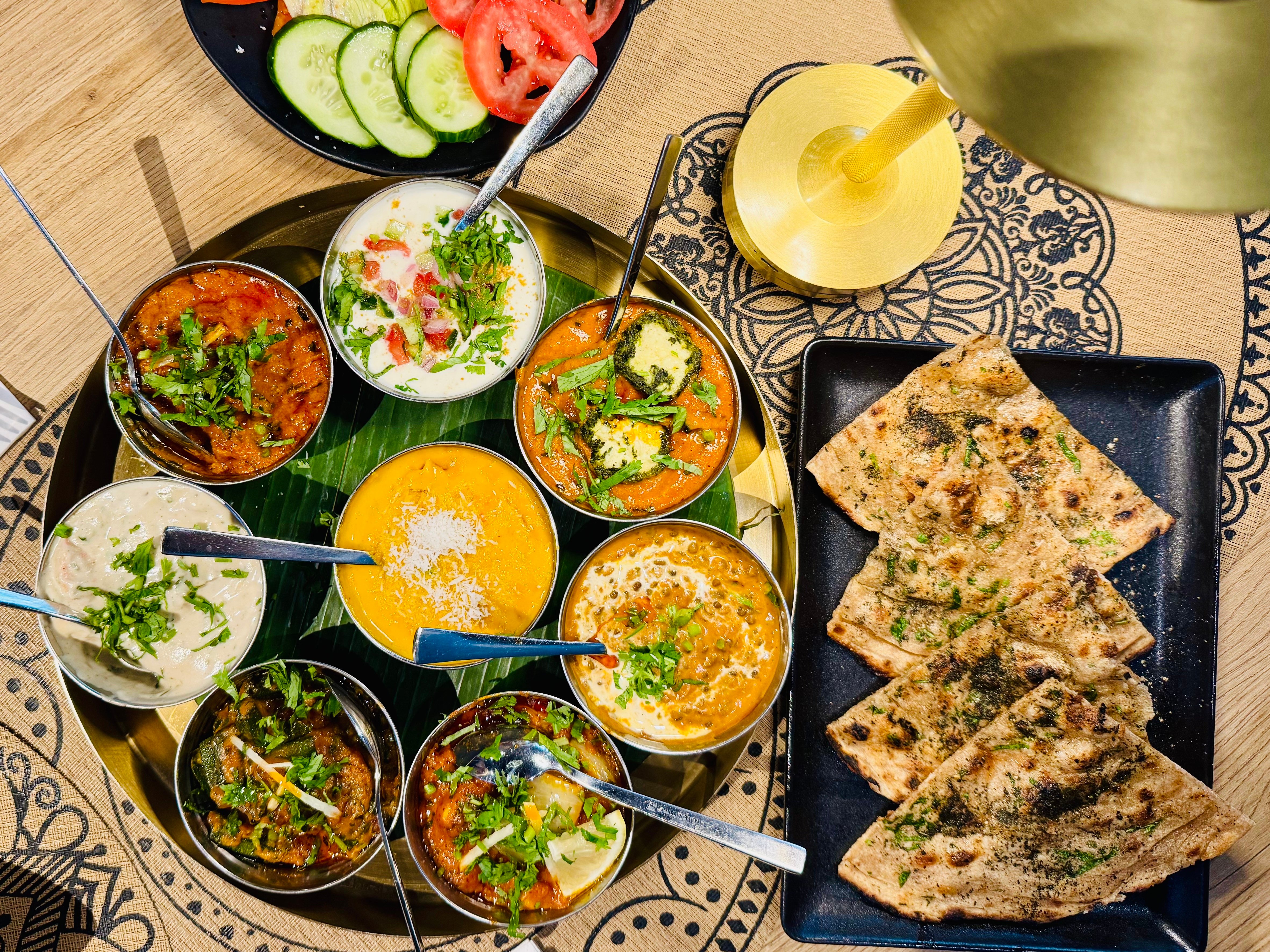 Rundes Thali-Tablett mit mehreren Schälchen Curry, Daal, Raita und Salat, daneben Fladenbrot