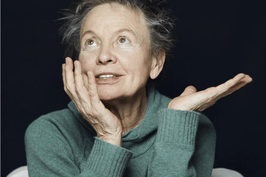 Older woman in a teal sweater gazing upward with a thoughtful smile, hands near her face. (informative portrait).