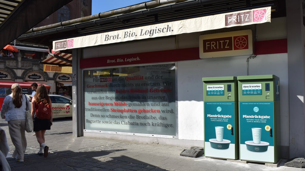 Shopfront with FRITZ bakery sign reading 'Brot. Bio. Logisch.' and two people walking by on a sunny street.