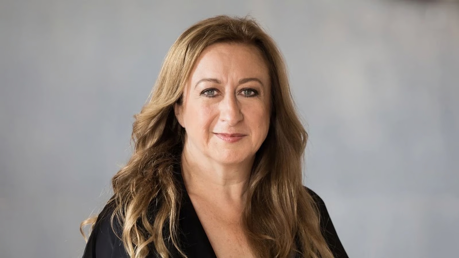 Professional headshot of a smiling woman with long wavy light brown hair, wearing a black top, against a neutral gray backdrop.