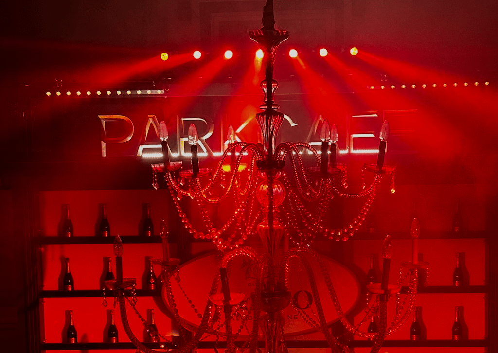 Red-lit bar interior with a crystal chandelier in the foreground and bottle shelves in the background.