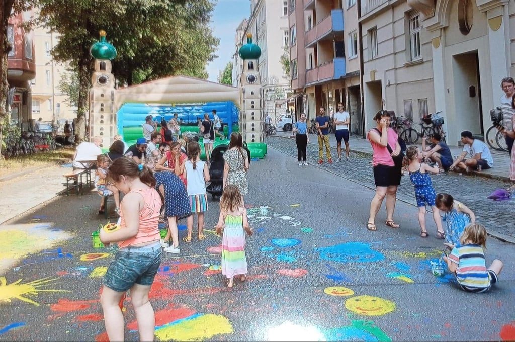 Children drawing with bright sidewalk chalk on a sunlit street while adults watch nearby; a blue inflatable structure is in the background.