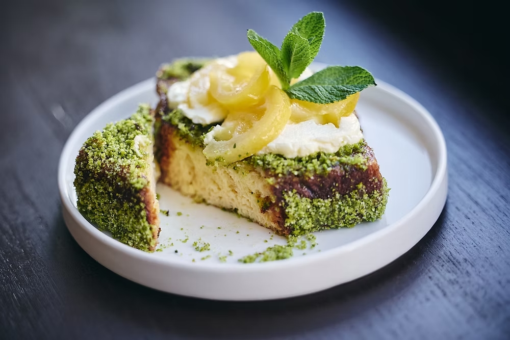 Slice of pistachio-crusted cake with cream frosting, lemon slices, and mint on a white plate.