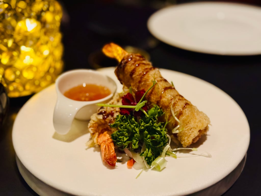 Fried shrimp atop greens with a small cup of dipping sauce on a white plate, gold decor blurred in the background.