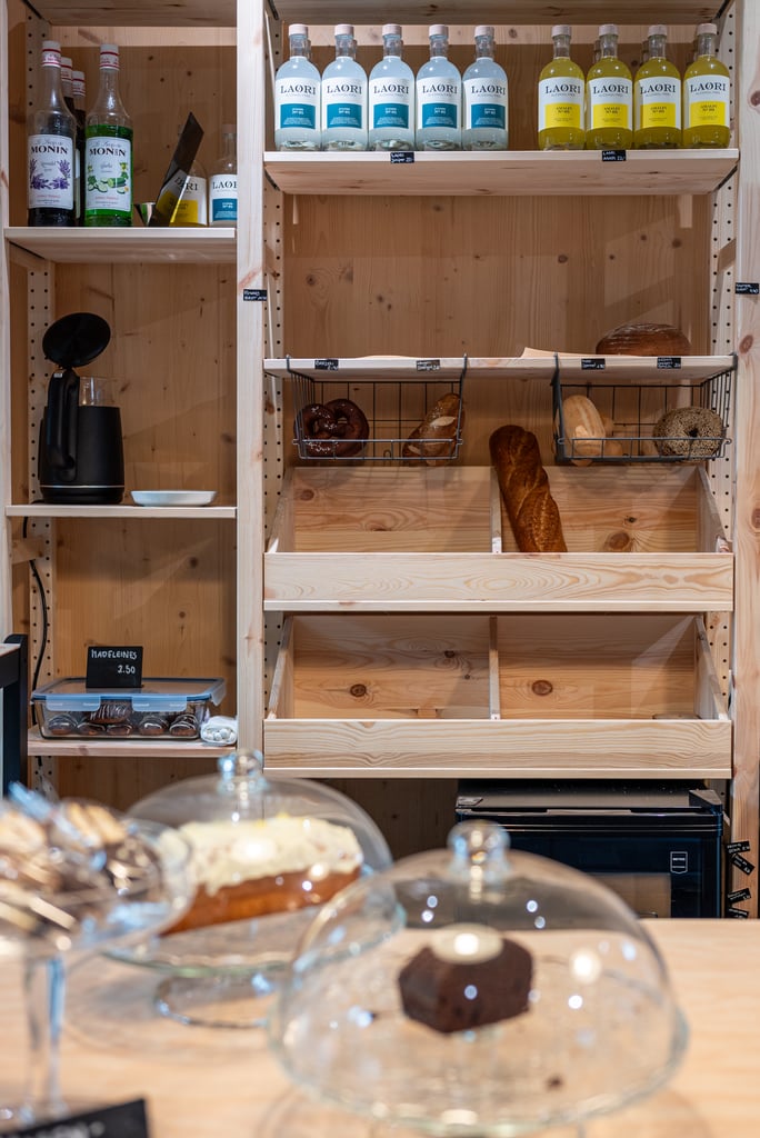 Wooden bakery display with bottles on the top shelf, assorted breads and pretzels on lower shelves, and glass cake domes in the foreground