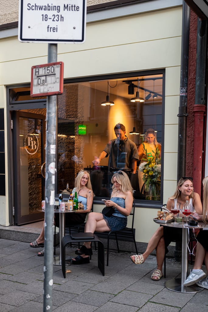 Group of friends dining outside a city cafe; three blond women sit at a table with drinks and food, a man and woman inside visible through the window.