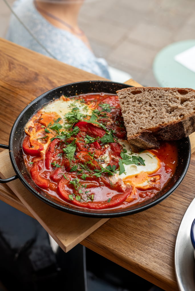 Skillet of shakshuka with poached eggs in tomato sauce, peppers, herbs, and a slice of crusty bread on the edge.