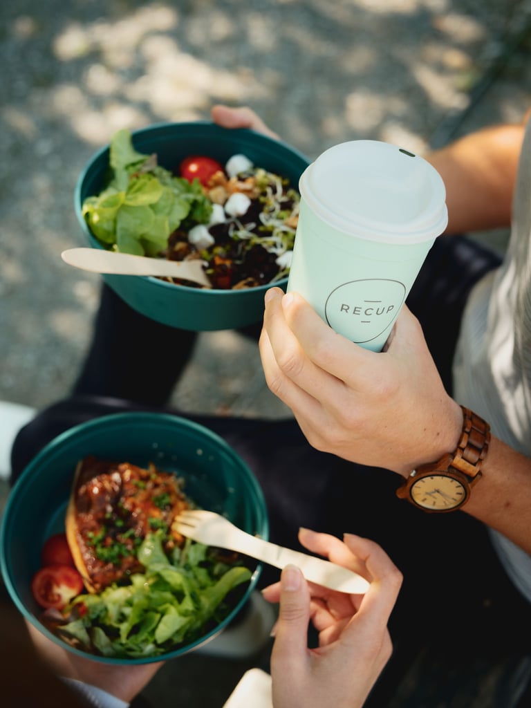 Person holds a light-blue takeaway cup while sharing a green salad bowl outdoors.