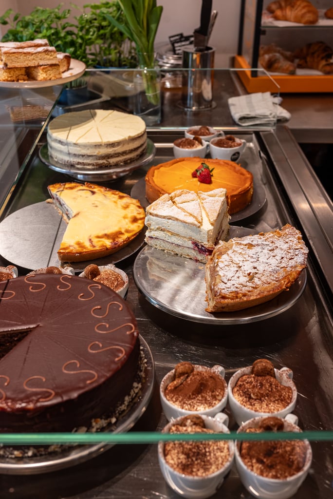 Assorted cakes and pastries displayed in a bakery case, including a chocolate frosted cake, orange cheesecake, and layered vanilla cake slices behind glass.