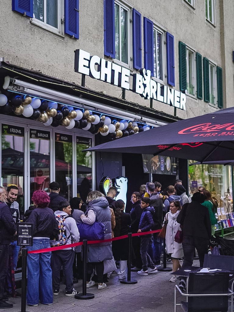 People line up outside a bar with blue window shutters and a bold sign reading ECHTE BERLINER, under a purple umbrella and festive balloons nearby, on a city street.
