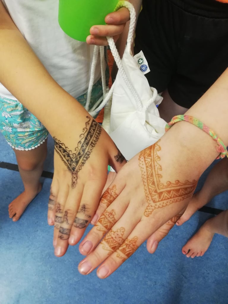 Group of hands with detailed henna designs on forearms and fingers, wearing bracelets, on a blue floor with a white bag and green cup nearby.