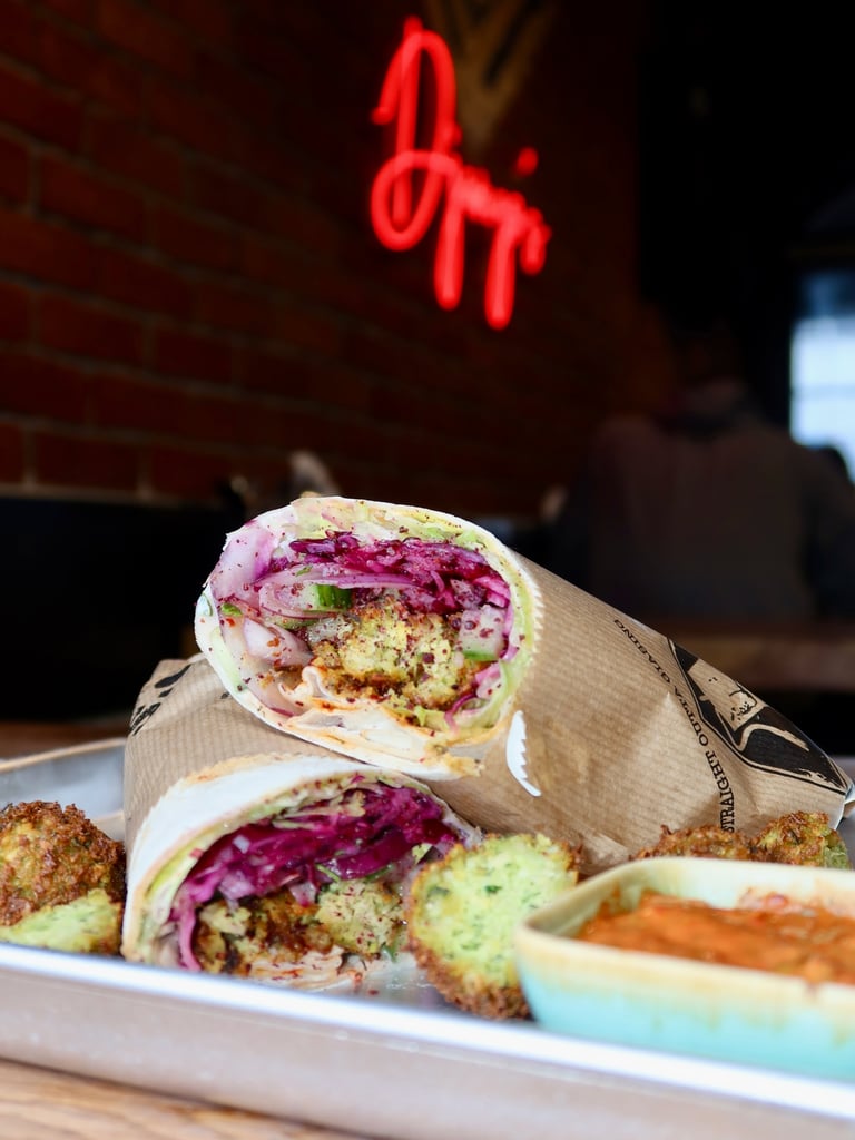 Close-up of a colorful veggie wrap cut in half on a tray, with a blurred red neon 'Open' sign in the background.