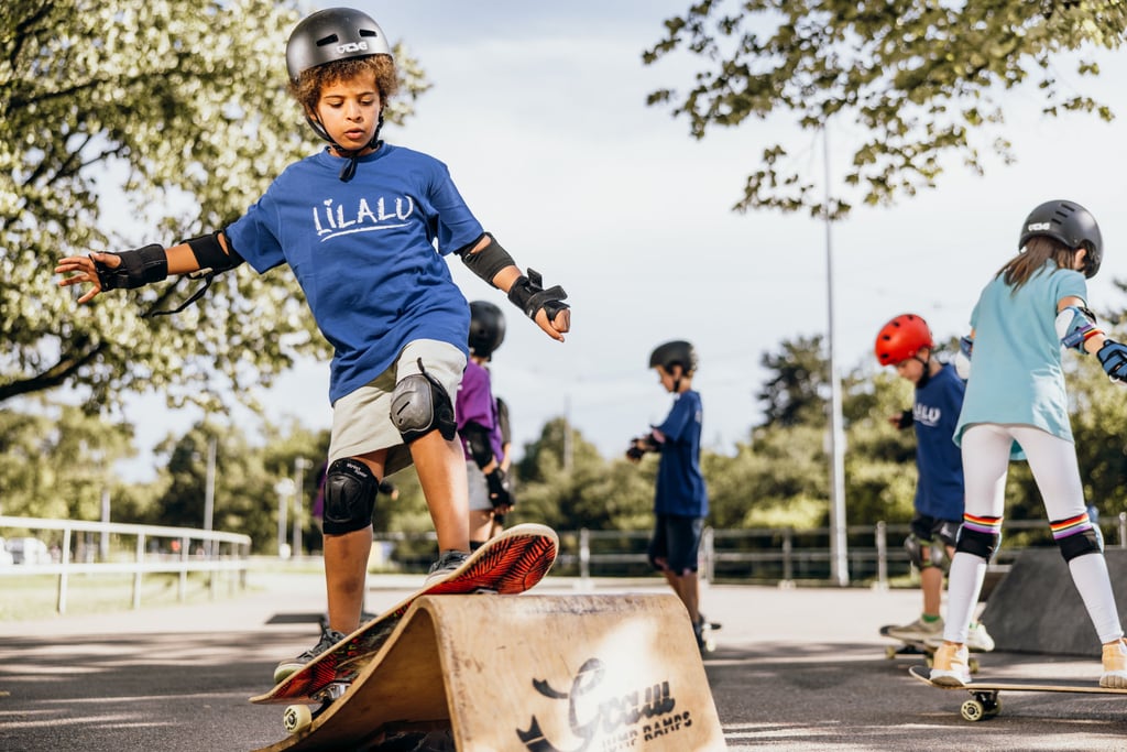 Child in a blue shirt and helmet jumping over a wooden skate ramp at an outdoor park with other skaters watching behind him, sunny day.