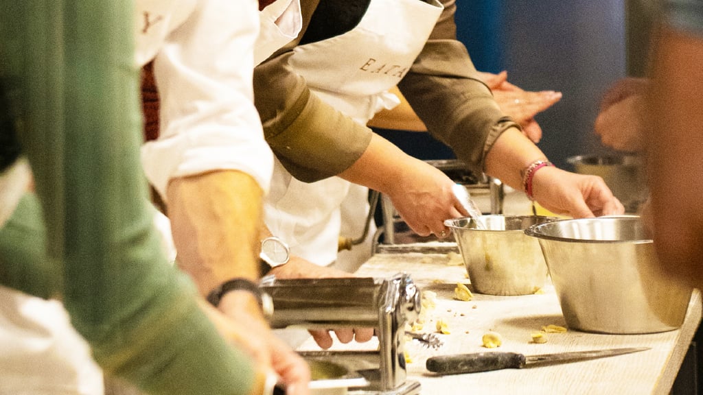 People kneading dough with a pasta machine and metal bowls on a kitchen counter, in a hands-on cooking class.