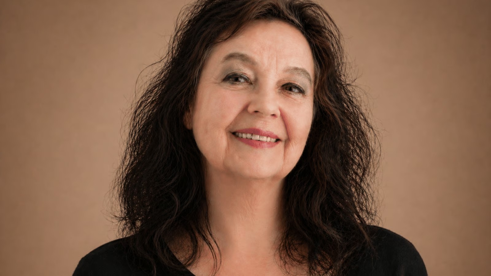 Smiling woman with shoulder-length dark curly hair against a brown studio backdrop.