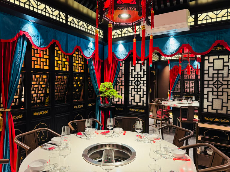 Elegant Chinese restaurant interior with black lattice walls, blue drapes, and red tassels; round tables with built-in grills ready for hotpot dining.