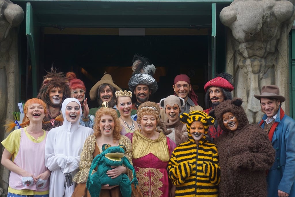 Group of performers in colorful costumes (princess, tiger, frog, bear, ghost) posing together at an entrance.