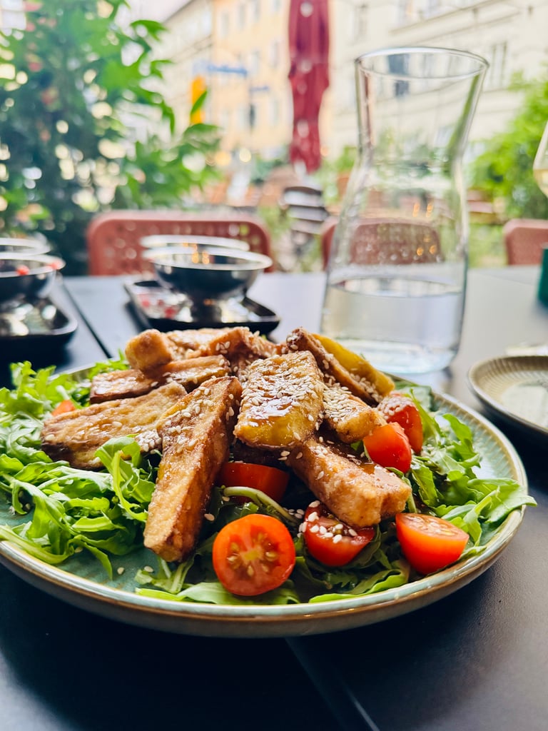 Plate of sesame-crusted fried breadsticks on a bed of lettuce and cherry tomatoes at an outdoor cafe table.Healthy greens on the side, blurred background.
