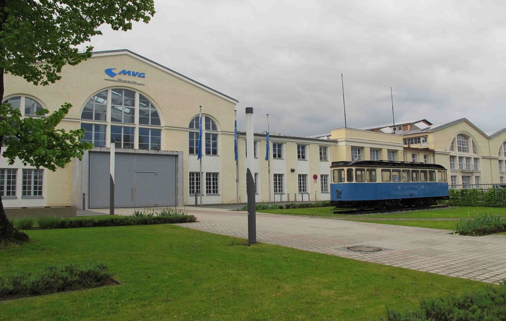 MVG Museum building with arched windows, a blue tram on tracks in front, and green lawn nearby
