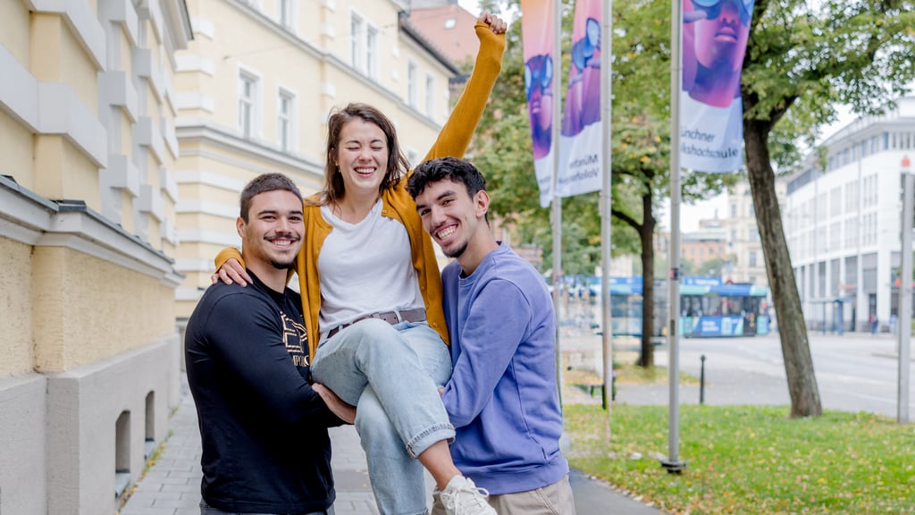 Three friends outdoors in an urban street; a woman in a mustard cardigan is lifted by two men, all smiling.