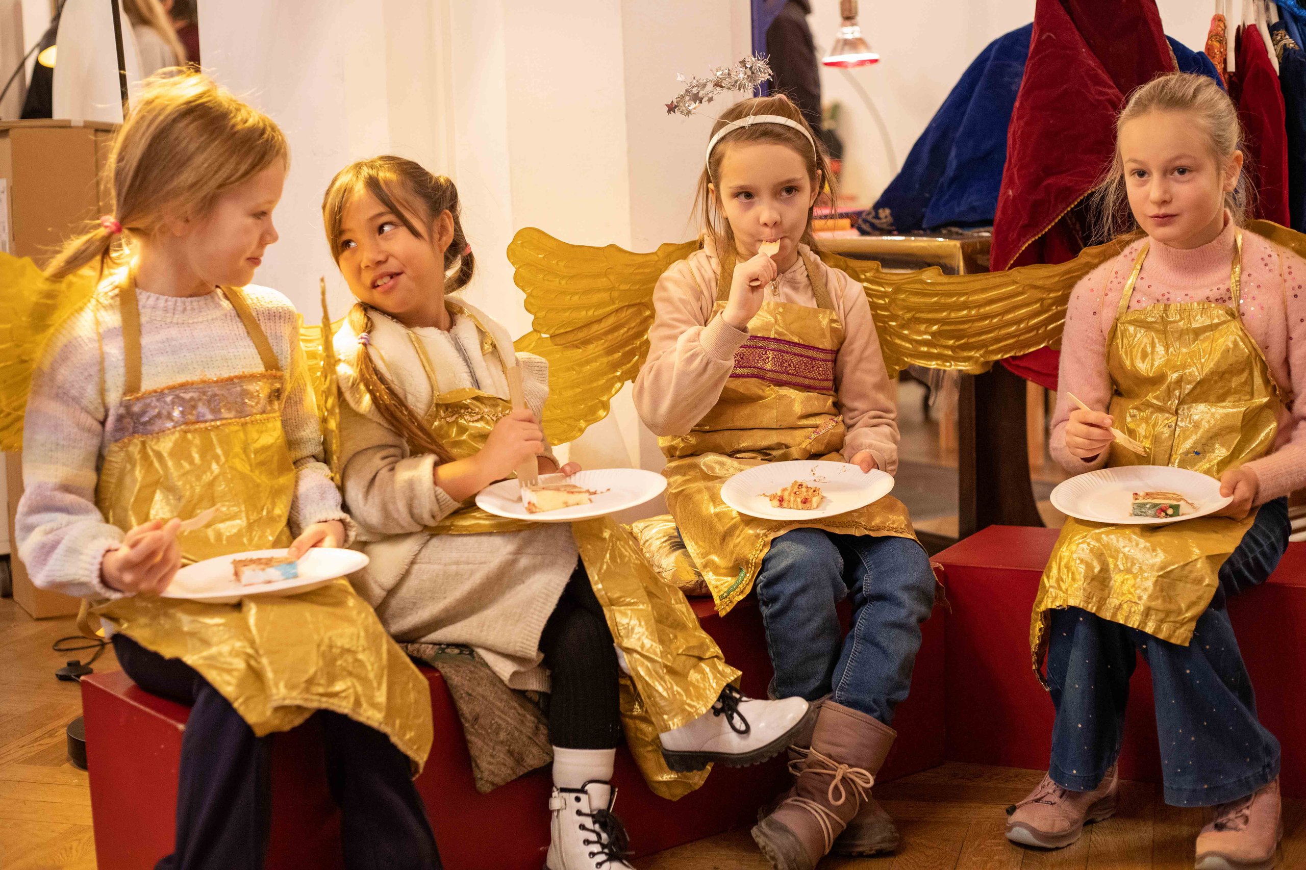 Vier Mädchen sitzen als Engel verkleidet in einer Reihe und essen Kuchen.