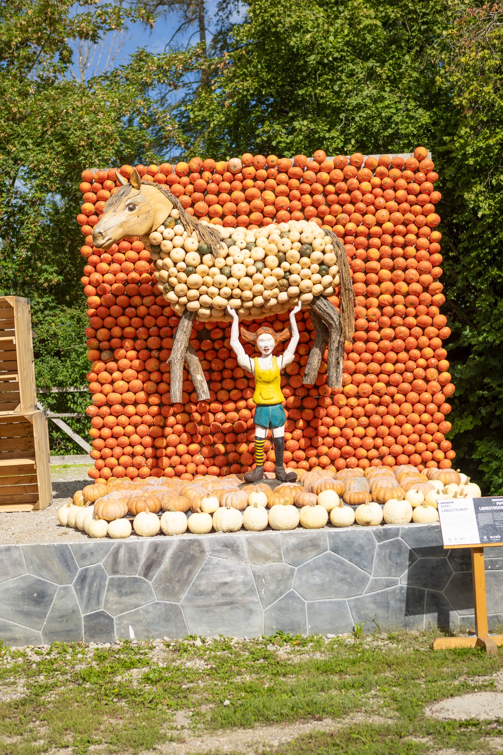 Outdoor pumpkin sculpture: a life-size horse made from pumpkins with a person figure beneath, in front of a wall of stacked orange pumpkins and a circular pumpkin base base.