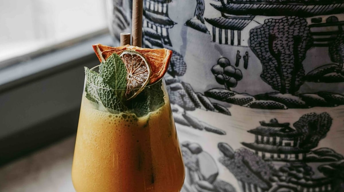 Cocktail mit getrockneter Orangenscheibe und Minze vor einer Vase mit blau-weißem Muster