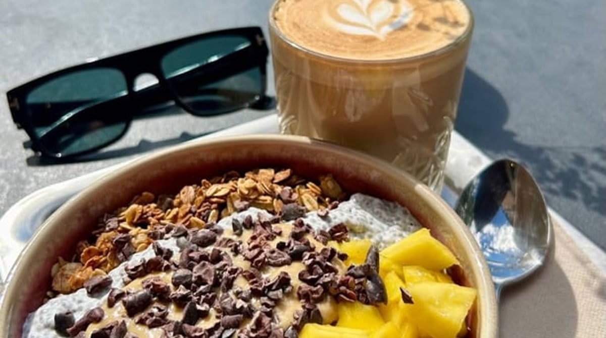Açaí-Bowl mit Heidelbeeren, Mango, Granola und Schokolade, dahinter ein Cappuccino