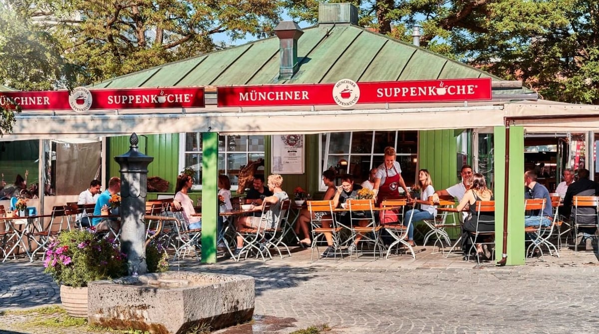 Die Münchner Suppenküche auf dem Viktualienmarkt mit grünem Dach, Biergartentischen und Gästen im Freien