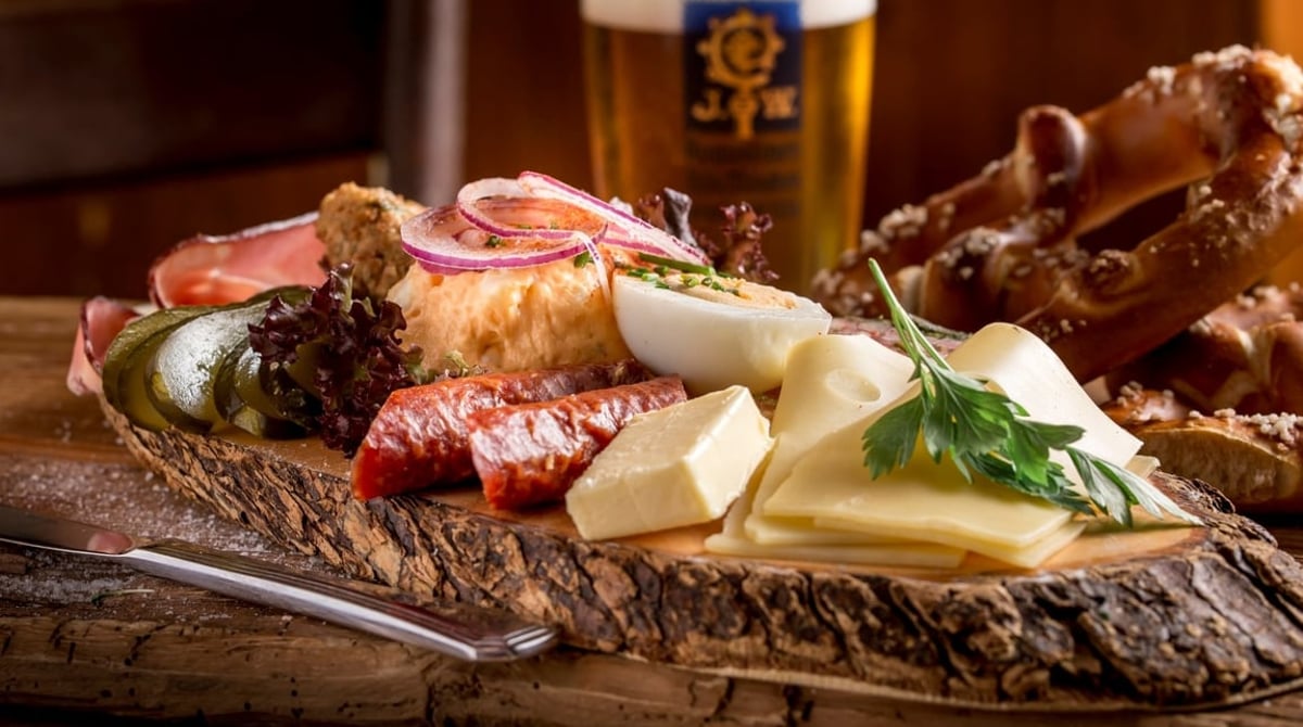 Brotzeitbrett mit Wurst, Käse und Brezn, dahinter ein Glas Augustiner