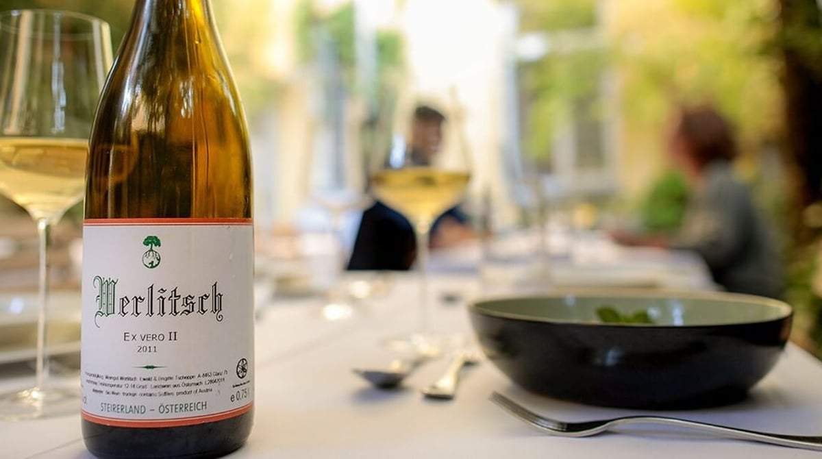 Gedeckter Tisch mit einer Flasche Weißwein und Weingläsern, im Hintergrund Grün