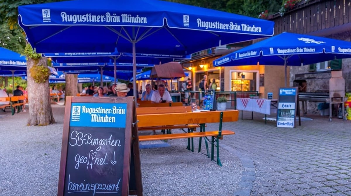 Biergartentische und blaue Augustiner-Sonnenschirme unter Kastanien im Kastanienbiergarten des Gasthofs Hinterbrühl, davor eine Tafel „SB-Biergarten geöffnet“