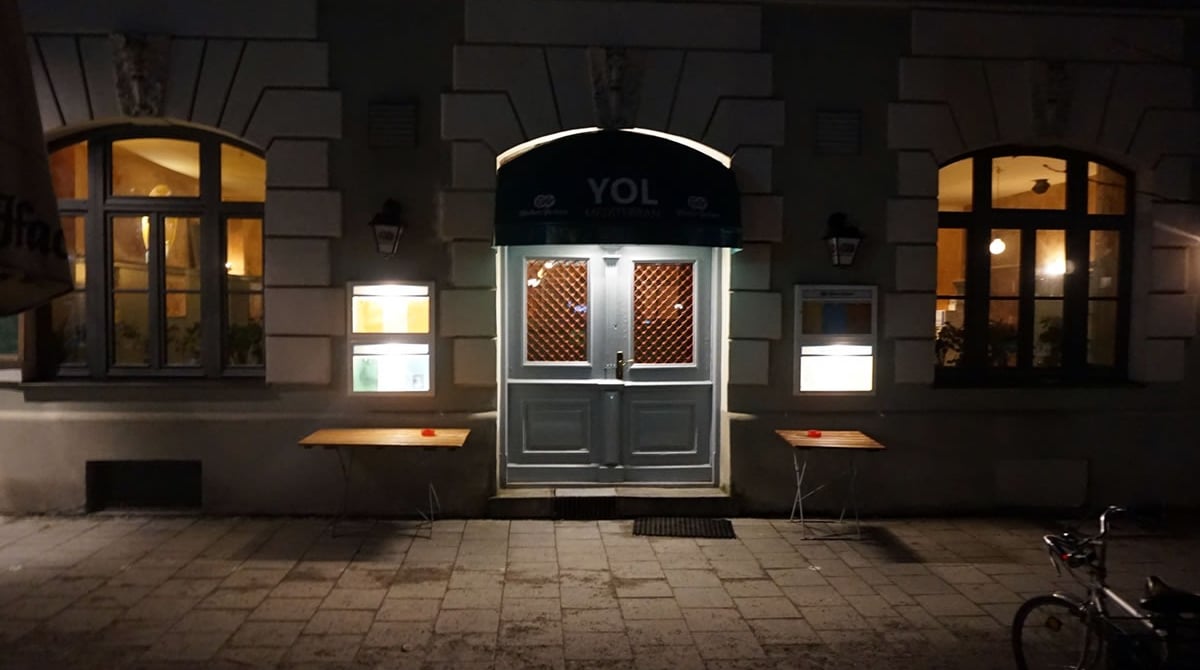 Beleuchteter Eingang der Yol Taverne in München am Abend, davor zwei Tische