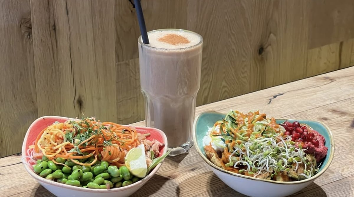 Zwei bunte Bowls mit Edamame, Zoodles und Granatapfel und ein Smoothie auf einem Holztisch bei Bowls & Blenders