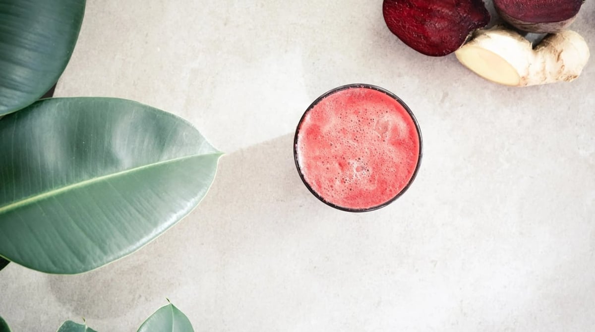 Glas mit rotem Rote-Bete-Saft auf hellem Tisch, daneben Rote Bete und Ingwer