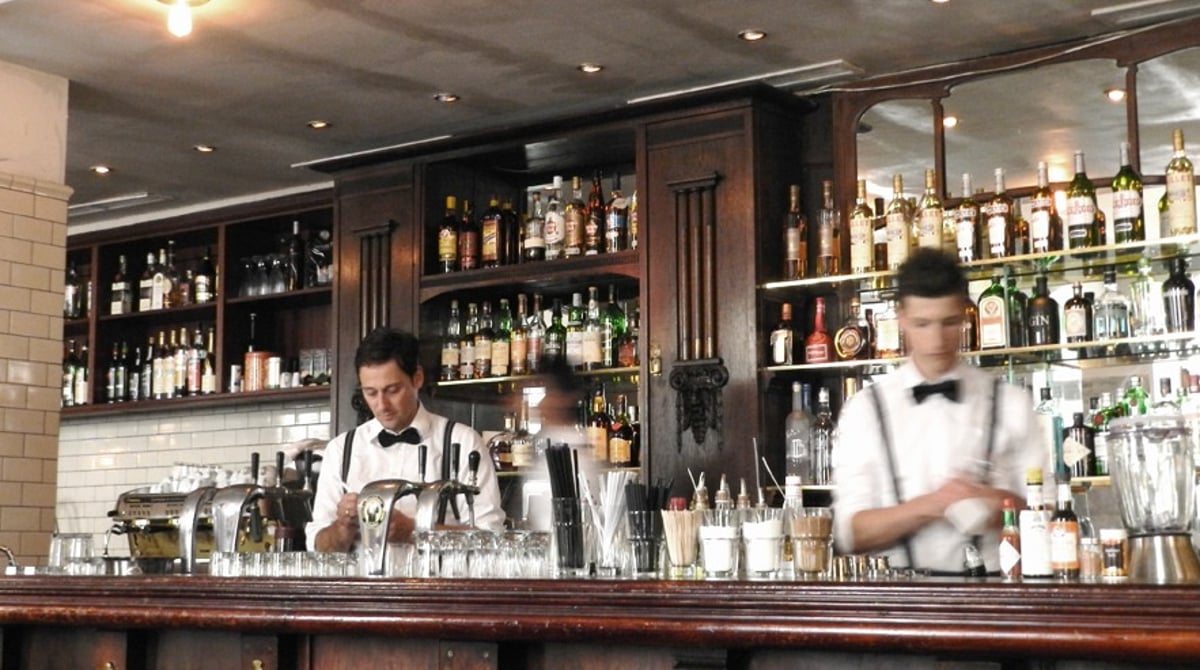 Dunkle Holzbar des Chez Fritz mit Flaschenregal und weißen Kacheln, dahinter zwei Barkeeper mit Fliege