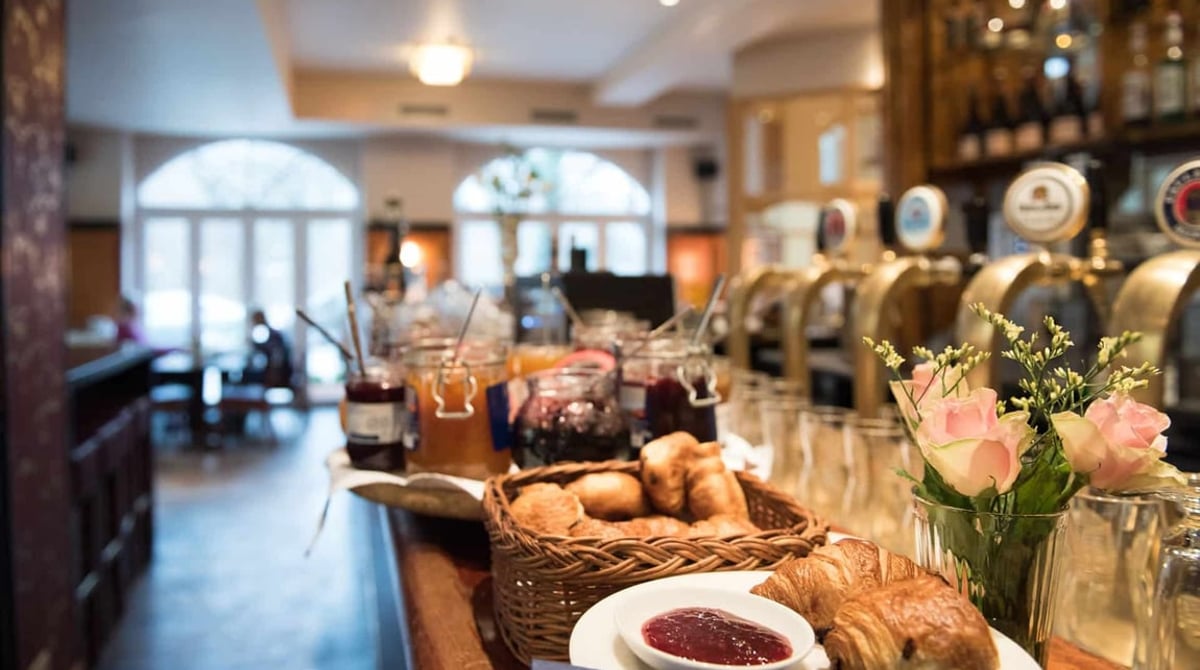 Gastraum des Hotel Ritzi mit Holzbar und Zapfhähnen, davor Croissants und Marmeladen auf dem Frühstücksbuffet, im Hintergrund hohe Bogenfenster