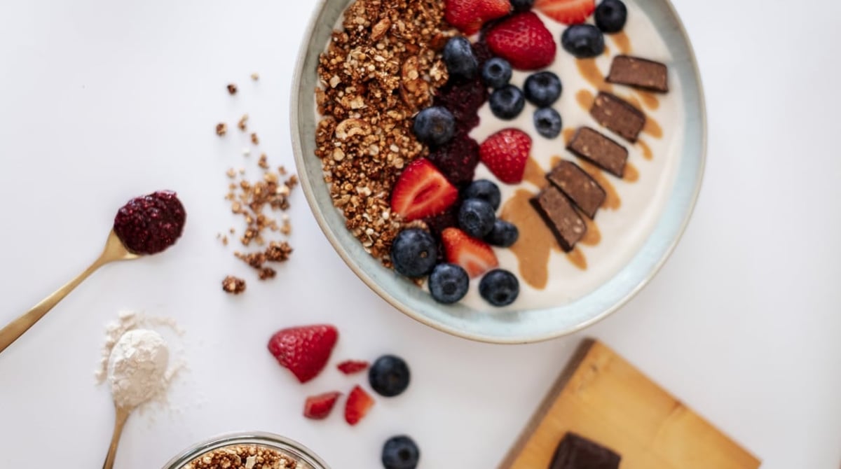 Bowl mit Granola, Erdbeeren, Heidelbeeren und Schokoriegel-Stücken auf Joghurt, daneben Löffel mit Beerenpulver und Mehl