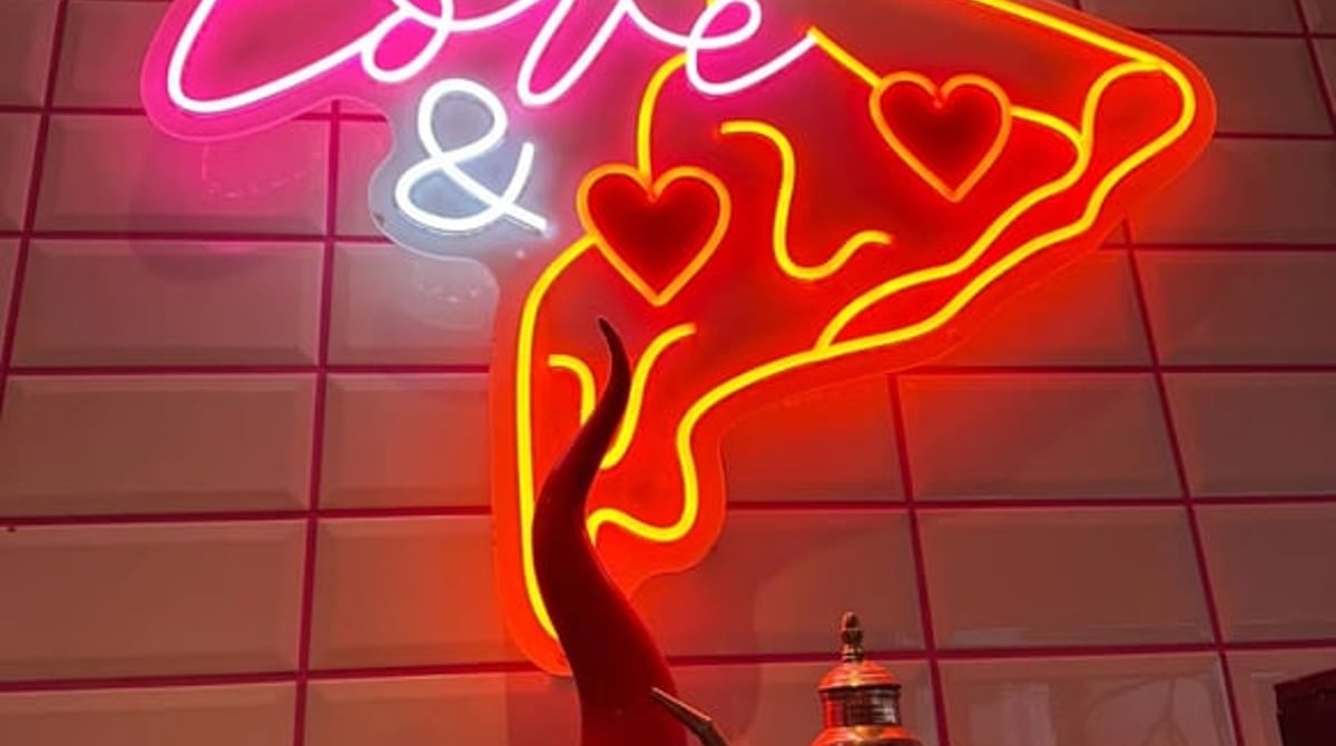 Neon-Schild „All you need is love" mit Pizza-Motiv auf rosa Fliesenwand