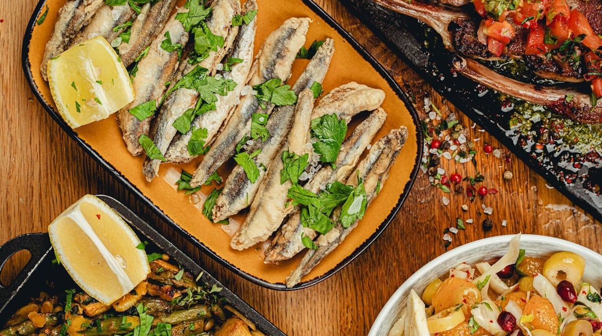 Gebratene Boquerones auf einer gelben Platte, umgeben von weiteren Tapas auf einem Holztisch im Amistad