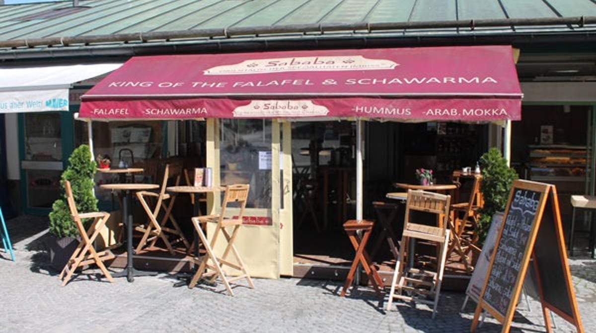 Außenansicht des Sababa auf dem Viktualienmarkt mit roter Markise, Tischen und Kreidetafel