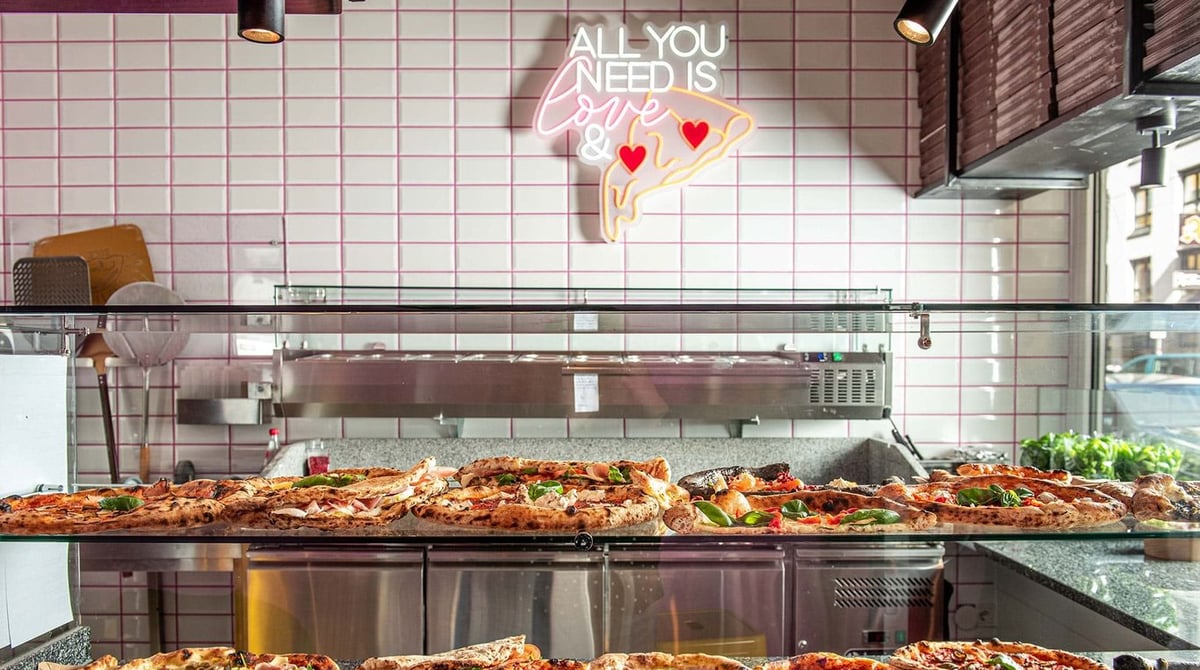 Pizzastücke in der Glasvitrine vor einer gefliesten Wand mit Neon-Schild im Napoli Slice