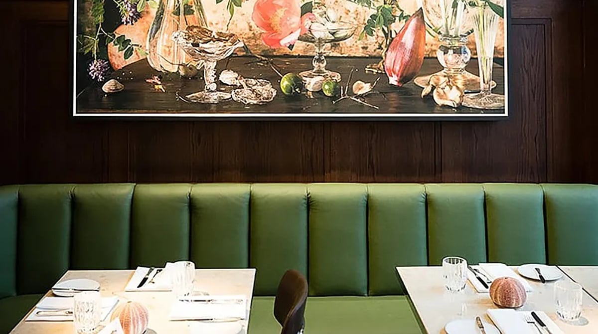 Gastraum der Dallmayr Bar & Grill mit grüner Lederbank, gedeckten Marmortischen und großem Stillleben-Gemälde an der Holzwand